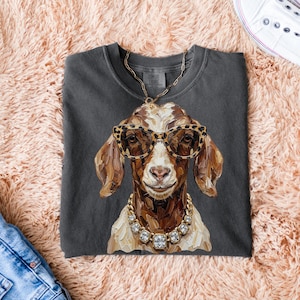 May include: A charcoal gray crewneck t-shirt featuring a painterly illustration of a goat wearing leopard print glasses and a chunky gold-toned rhinestone necklace. The goat has brown and white fur, and the shirt has a visible size M tag.