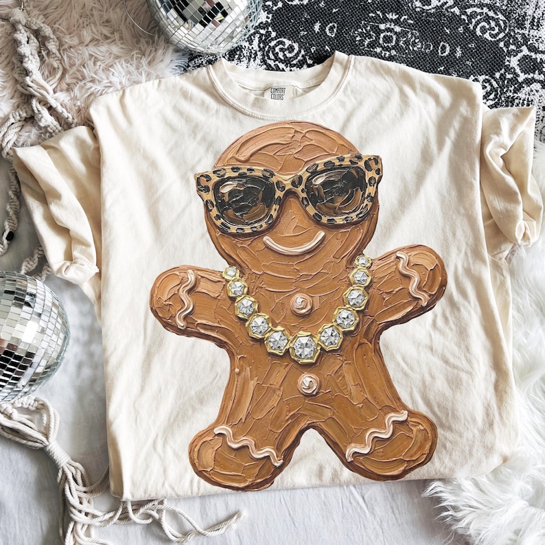 May include: A cream-colored t-shirt featuring a central graphic of a brown gingerbread person. The gingerbread figure wears leopard print sunglasses and a gold-toned chain necklace with hexagonal links. The design has a textured, painted appearance.