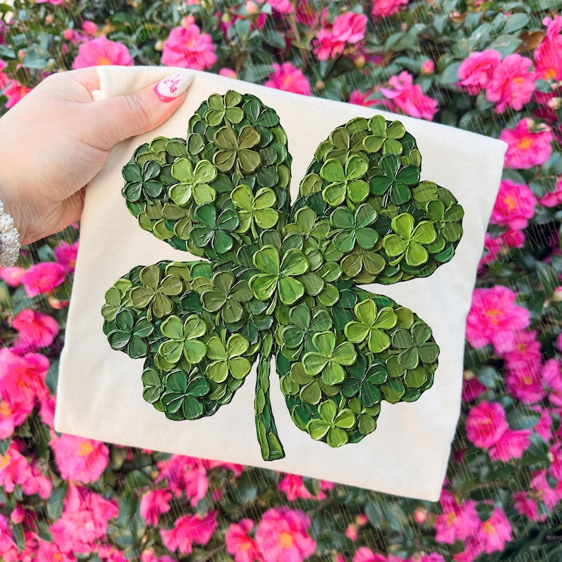 May include: A square, off-white canvas features a large, textured four-leaf clover design. The clover is composed of many small, green clovers, with varying shades of green. The background is a blur of pink flowers.