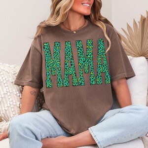 K&ouml;nnte beinhalten: Ein braunes T-Shirt mit kurzen &Auml;rmeln und dem Wort MAMA in gro&szlig;en Blockbuchstaben. Die Buchstaben haben ein leuchtend gr&uuml;nes und schwarzes Leopardenmuster. Das T-Shirt hat einen Rundhalsausschnitt und eine lockere Passform aus braunem Stoff.