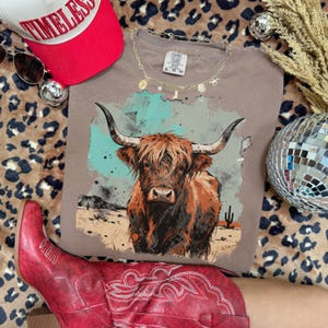 May include: A taupe t-shirt with a watercolour illustration of a Highland cow. The shirt is styled with a red baseball cap, sunglasses, and red cowboy boots. The shirt is size XL-TG. The background includes a leopard print and a disco ball.