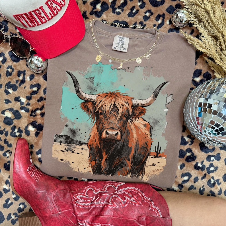 May include: A taupe t-shirt with a watercolour illustration of a Highland cow. The shirt is styled with a red baseball cap, sunglasses, and red cowboy boots. The shirt is size XL-TG. The background includes a leopard print and a disco ball.