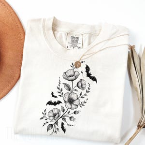 May include: Cream-colored t-shirt featuring a floral and bat design. The graphic includes black and white flowers, leaves, and bats. A gold necklace with a pendant is draped over the shirt. The shirt has a Comfort Colors tag.