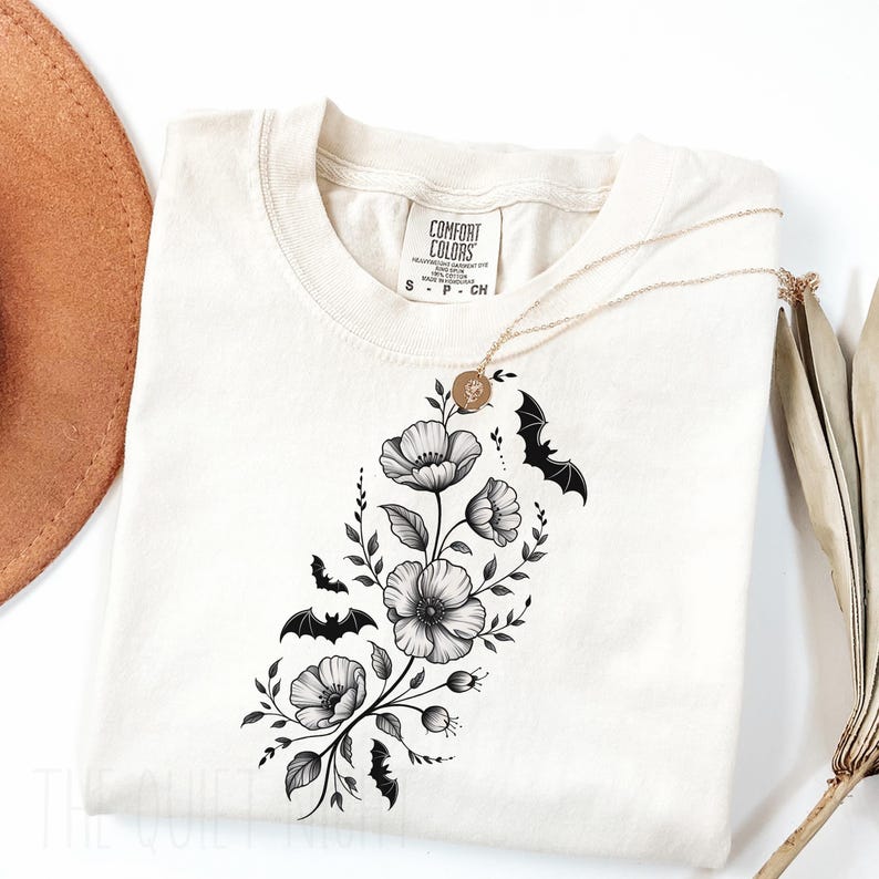May include: Cream-colored t-shirt featuring a floral and bat design. The graphic includes black and white flowers, leaves, and bats. A gold necklace with a pendant is draped over the shirt. The shirt has a Comfort Colors tag.