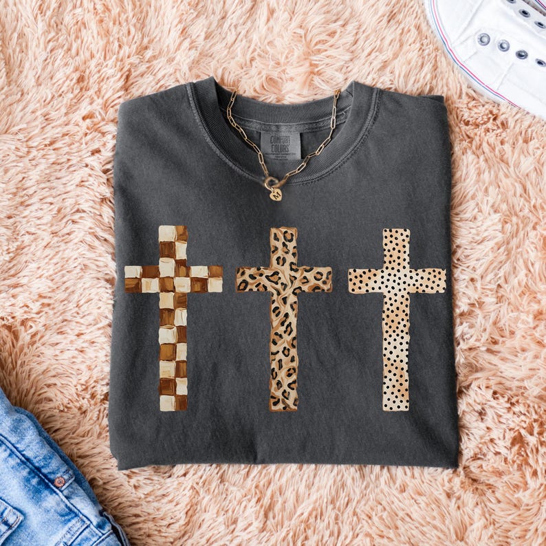 May include: Dark gray t-shirt featuring three crosses with unique patterns: a checkered design, a leopard print, and a dotted pattern. A gold chain necklace with a pendant is visible. The shirt is folded on a pink, fluffy surface.