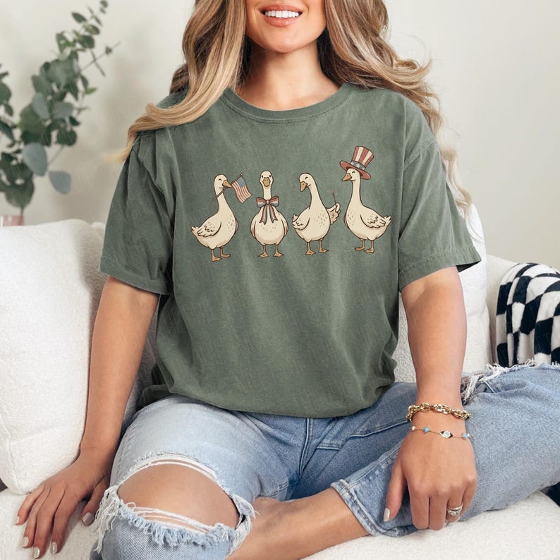 May include: Olive green t-shirt featuring a whimsical illustration of four geese. One goose holds a small American flag, another wears a bow tie, and the last wears a top hat. The geese are in a line.