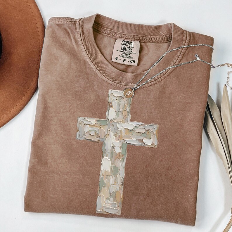 May include: A brown Comfort Colors t-shirt with a large, textured cross design in shades of white, green, and beige. A silver necklace with a small pendant rests on the shirt. The shirt is size S-P-CH.