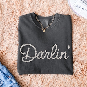 Pu&ograve; includere: T-shirt grigio scuro con la scritta "Darlin'" in perle bianche. Una collana dorata con un ciondolo &egrave; appoggiata sulla maglietta. La maglietta &egrave; piegata su una superficie rosa soffice, con un paio di jeans blu e una scarpa bianca visibili.