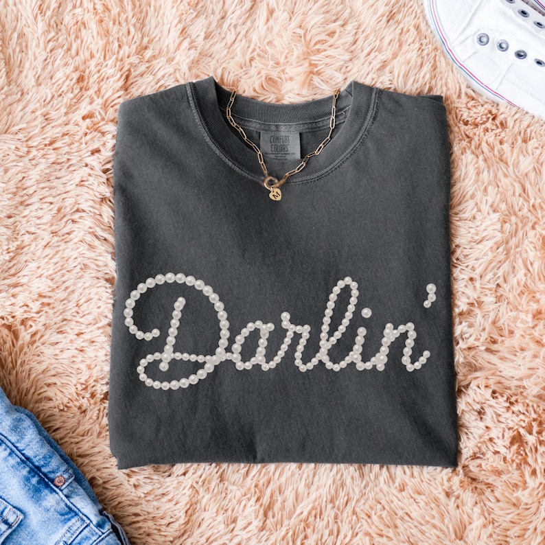 Pu&ograve; includere: T-shirt grigio scuro con la scritta "Darlin'" in perle bianche. Una collana dorata con un ciondolo &egrave; appoggiata sulla maglietta. La maglietta &egrave; piegata su una superficie rosa soffice, con un paio di jeans blu e una scarpa bianca visibili.