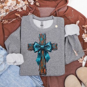 Peut inclure: Un sweat-shirt gris avec un motif de croix peinte et un n&oelig;ud bleu sarcelle. La croix a un aspect textur&eacute; et rustique avec des tons marron et sarcelle. Le sweat-shirt est de style col rond.