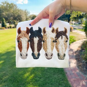 Peut inclure: Sweat-shirt blanc orn&eacute; de quatre t&ecirc;tes de chevaux brod&eacute;es en marron, noir et beige. Les t&ecirc;tes de chevaux sont d&eacute;taill&eacute;es avec des coutures textur&eacute;es, mettant en valeur leurs crini&egrave;res et leurs traits. Le sweat-shirt est tenu par une personne.
