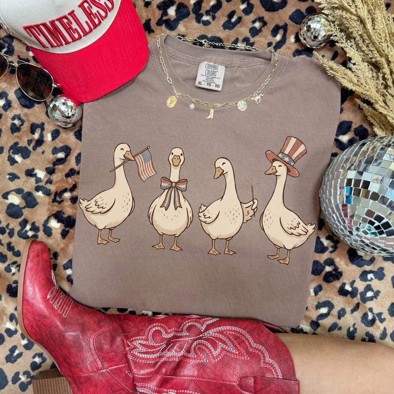 May include: A brown t-shirt featuring four cartoon geese, each adorned with patriotic accessories like an American flag, bow tie, and Uncle Sam hat. The shirt is paired with a red hat and boots, and a gold necklace.