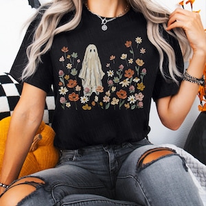 Może przedstawiać: Czarny t-shirt z kr&oacute;tkim rękawem z centralną grafiką przedstawiającą białego ducha otoczonego r&oacute;żnorodnymi kolorowymi polnymi kwiatami w odcieniach pomarańczu, ż&oacute;łci, r&oacute;żu i zieleni. Wz&oacute;r jest nadrukowany na przodzie bawełnianej tkaniny.