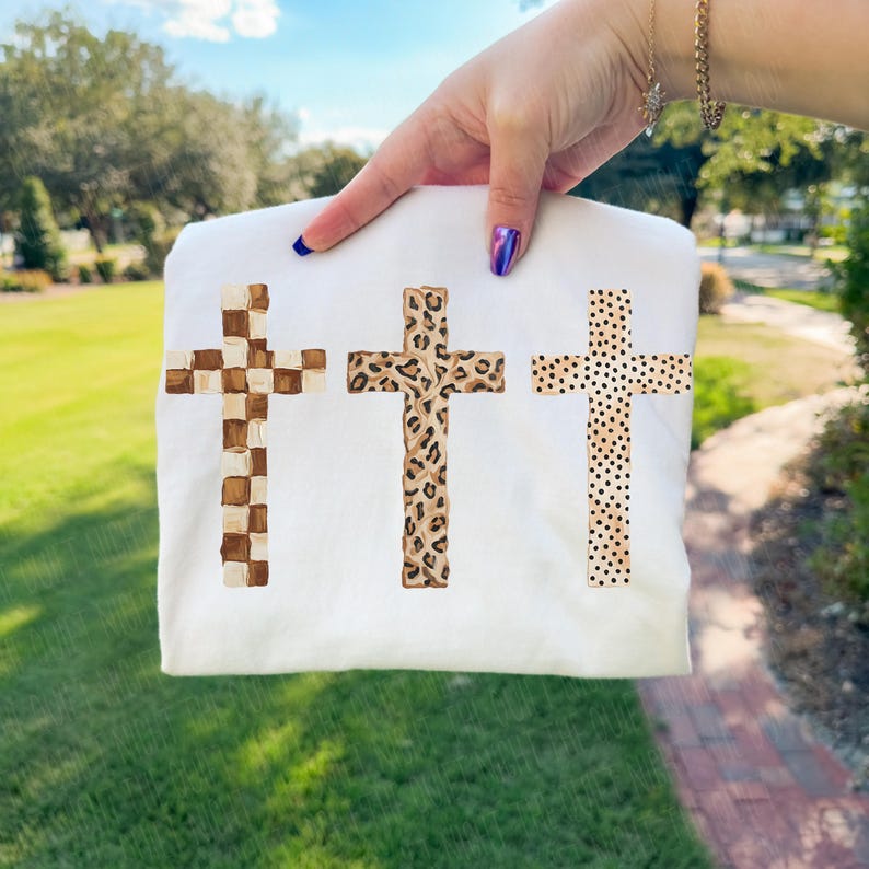 May include: White t-shirt featuring three crosses with unique designs. The crosses include a checkered pattern, a leopard print, and a dotted pattern. The shirt is held up by a person's hand, showcasing the design against a background of green grass and trees.