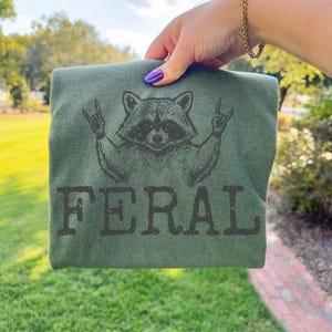May include: Green t-shirt featuring a raccoon graphic with the text "FERAL". The raccoon is depicted with its arms raised in a rock-and-roll gesture. The shirt is held up against a background of green grass and trees.