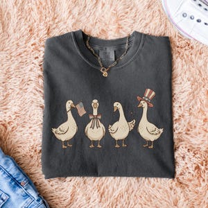 May include: Dark gray t-shirt featuring a whimsical illustration of four geese. Each goose is adorned with patriotic accessories: an American flag, a bow, a sparkler, and a top hat. The shirt is a Comfort Colors brand.