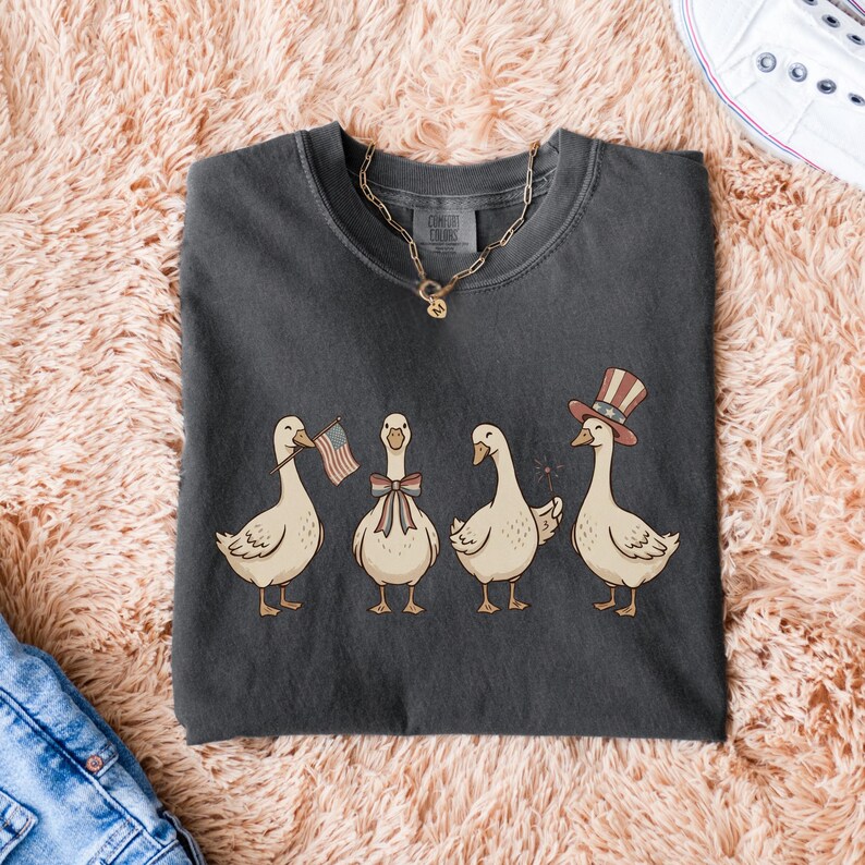 May include: Dark gray t-shirt featuring a whimsical illustration of four geese. Each goose is adorned with patriotic accessories: an American flag, a bow, a sparkler, and a top hat. The shirt is a Comfort Colors brand.