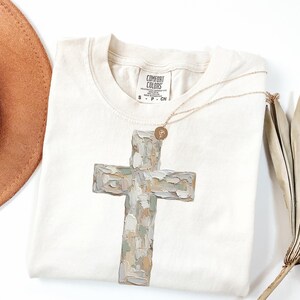 May include: Cream-colored t-shirt featuring a large, textured cross design in shades of gray, beige, and green. A gold necklace with a small pendant rests on the shirt. The shirt has a Comfort Colors label.
