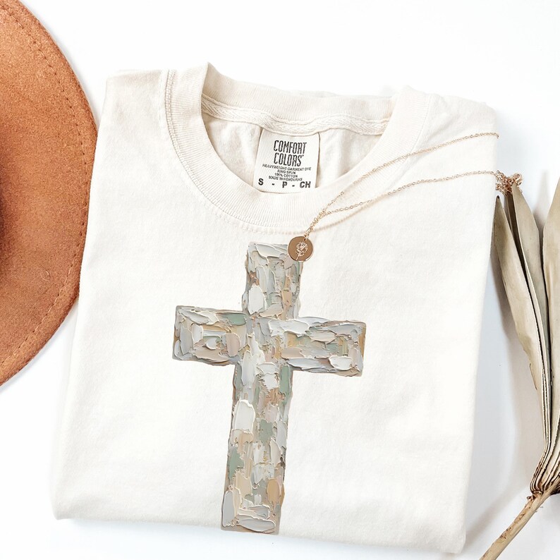 May include: Cream-colored t-shirt featuring a large, textured cross design in shades of gray, beige, and green. A gold necklace with a small pendant rests on the shirt. The shirt has a Comfort Colors label.