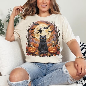 May include: A cream-colored t-shirt featuring a graphic print of a black cat sitting in front of a large yellow moon. The scene includes orange jack-o'-lanterns, bare trees, flying bats, and a silhouette of a witch on a broomstick against the moon.