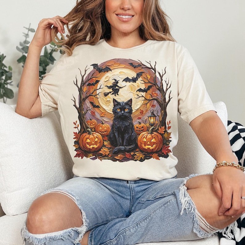 May include: A cream-colored t-shirt featuring a graphic print of a black cat sitting in front of a large yellow moon. The scene includes orange jack-o'-lanterns, bare trees, flying bats, and a silhouette of a witch on a broomstick against the moon.