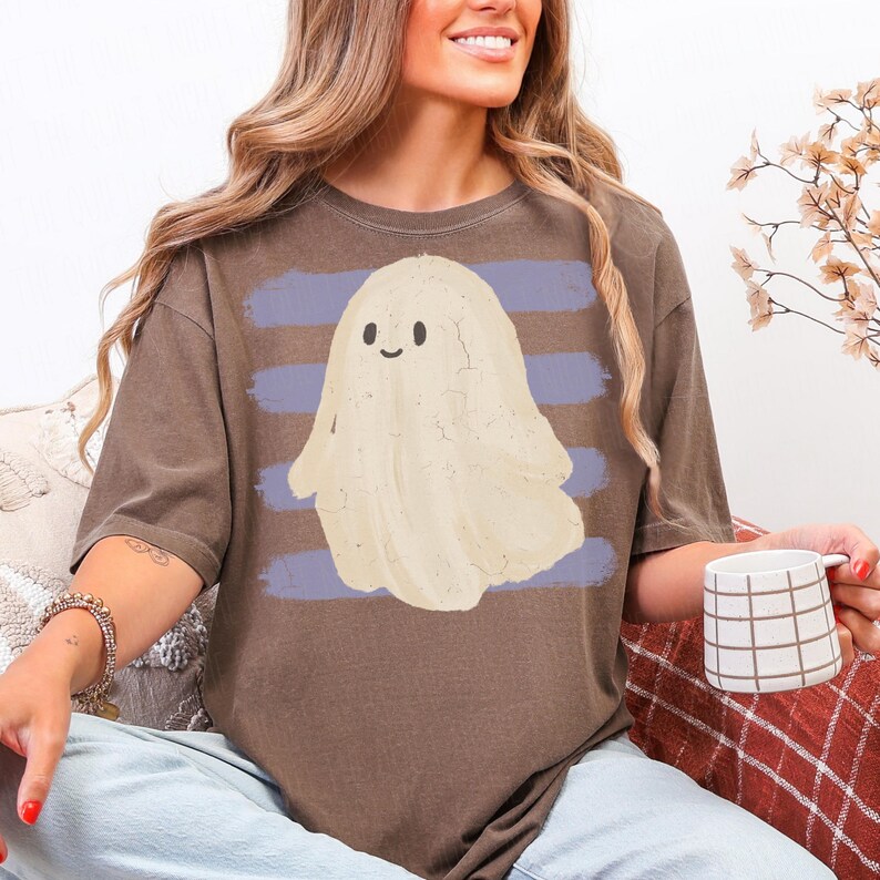 May include: A brown short-sleeve t-shirt featuring a graphic of a cream-colored ghost with a simple smiling face. The ghost is positioned over a background of horizontal light purple brushstroke stripes across the center of the garment.