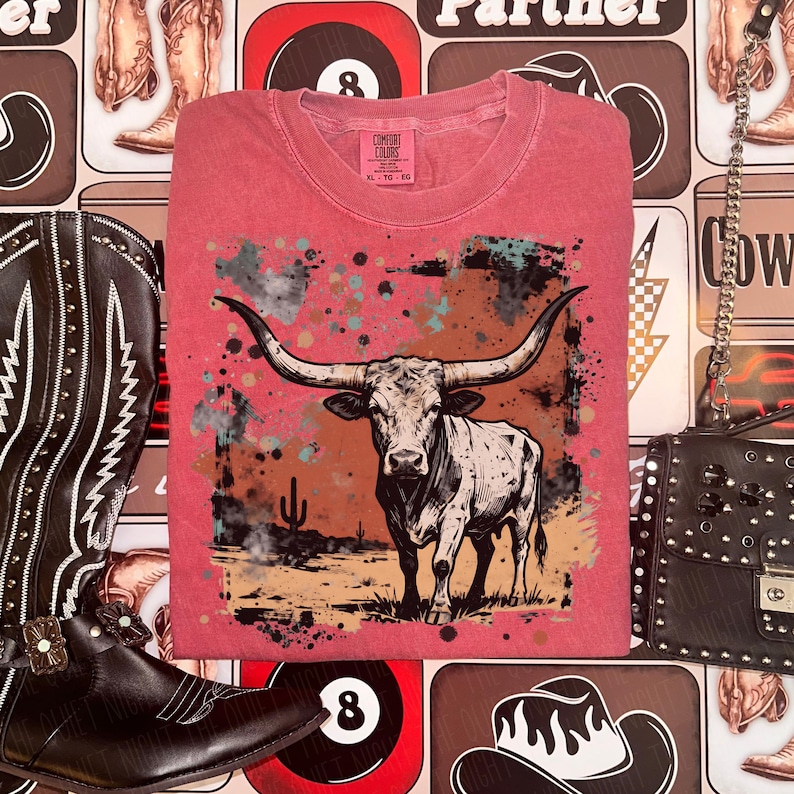 May include: A red t-shirt featuring a graphic of a longhorn steer in a desert landscape. The steer is white and black with large horns. The shirt has a Comfort Colors tag.