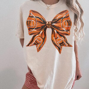 May include: Cream-colored t-shirt featuring a large, artistic bow design. The bow is painted in shades of orange, black, and white, resembling a basketball court. The shirt has short sleeves and a relaxed fit, suitable for casual wear.
