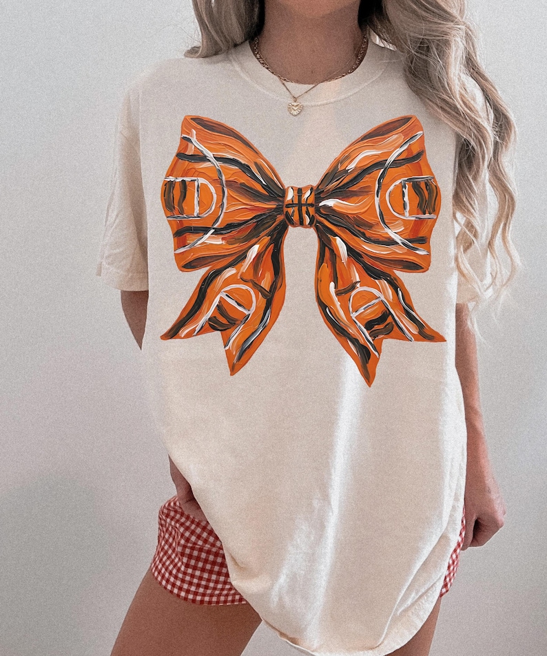 May include: Cream-colored t-shirt featuring a large, artistic bow design. The bow is painted in shades of orange, black, and white, resembling a basketball court. The shirt has short sleeves and a relaxed fit, suitable for casual wear.
