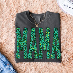 K&ouml;nnte beinhalten: Ein anthrazitfarbenes Sweatshirt mit Rundhalsausschnitt und dem Wort MAMA in gro&szlig;en Blockbuchstaben. Die Buchstaben haben ein leuchtend gr&uuml;nes und gelbes Leopardenmuster. Eine goldfarbene Kette liegt auf dem Kragen des Kleidungsst&uuml;cks.