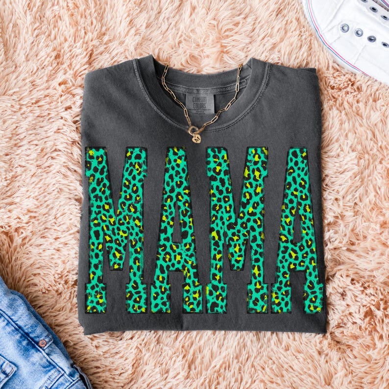 K&ouml;nnte beinhalten: Ein anthrazitfarbenes Sweatshirt mit Rundhalsausschnitt und dem Wort MAMA in gro&szlig;en Blockbuchstaben. Die Buchstaben haben ein leuchtend gr&uuml;nes und gelbes Leopardenmuster. Eine goldfarbene Kette liegt auf dem Kragen des Kleidungsst&uuml;cks.