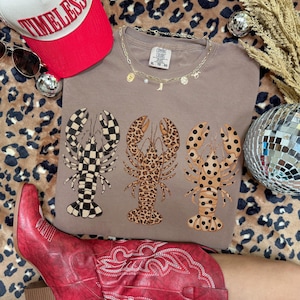 May include: A taupe Comfort Colors t-shirt featuring three lobster designs with different patterns: checkered, leopard print, and polka dots. The shirt is paired with a red baseball cap, sunglasses, and a gold necklace.