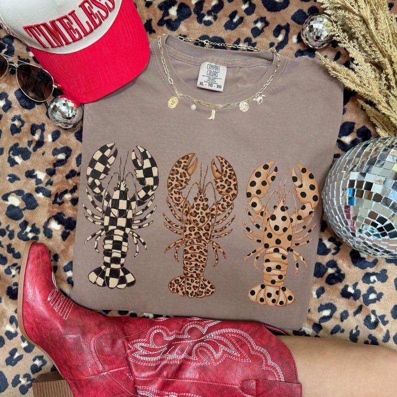 May include: A taupe Comfort Colors t-shirt featuring three lobster designs with different patterns: checkered, leopard print, and polka dots. The shirt is paired with a red baseball cap, sunglasses, and a gold necklace.