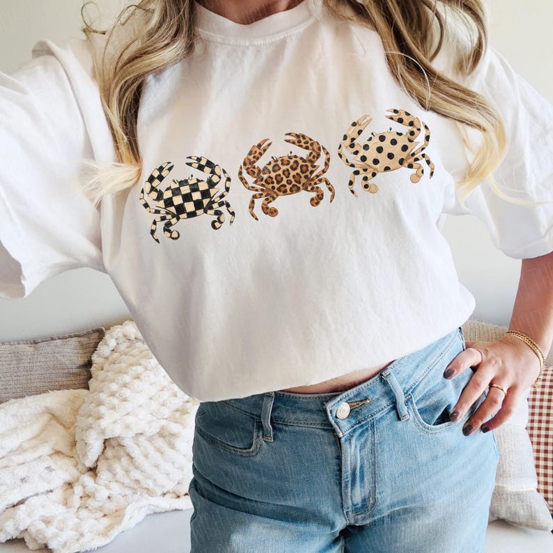 以下が含まれることがあります： 3匹のカニのデザインが施された白いTシャツ。カニは、白黒の市松模様、茶色のヒョウ柄、タンと黒のドット柄と、それぞれ異なる模様です。このシャツは柔らかい素材で作られています。