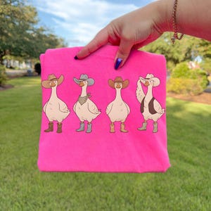 May include: A hot pink t-shirt featuring a cartoon design of four geese dressed as cowboys. Each goose wears a cowboy hat, boots, and one has a vest and bandana. The shirt is held up by a hand.