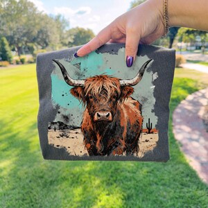 May include: A dark grey t-shirt with a watercolour illustration of a Highland cow. The cow has brown and orange fur, large horns, and a desert landscape background with cacti and a turquoise sky.