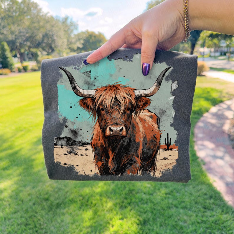 May include: A dark grey t-shirt with a watercolour illustration of a Highland cow. The cow has brown and orange fur, large horns, and a desert landscape background with cacti and a turquoise sky.