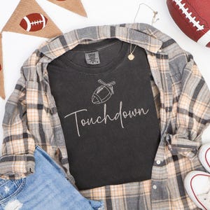 May include: A black t-shirt with the word "Touchdown" in cursive script and a football graphic. The shirt is layered with a plaid button-down shirt, blue jeans, and white sneakers. A football and a banner with a football are also in the image.