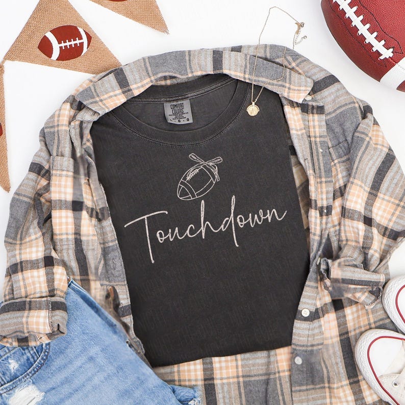 May include: A black t-shirt with the word "Touchdown" in cursive script and a football graphic. The shirt is layered with a plaid button-down shirt, blue jeans, and white sneakers. A football and a banner with a football are also in the image.