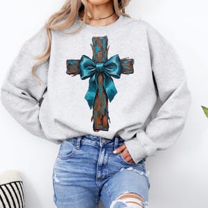Peut inclure: Un sweat-shirt gris clair avec une croix rustique. La croix est marron avec des accents turquoise et un n&oelig;ud turquoise. Le sweat-shirt est de style col rond et est fait d'une mati&egrave;re douce.