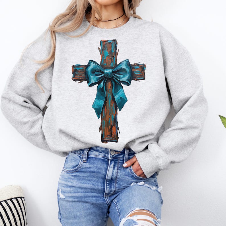 Peut inclure: Un sweat-shirt gris clair avec une croix rustique. La croix est marron avec des accents turquoise et un n&oelig;ud turquoise. Le sweat-shirt est de style col rond et est fait d'une mati&egrave;re douce.