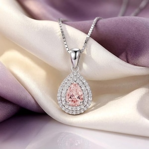 May include: A silver necklace with a pear-shaped pendant. The pendant features a light pink gemstone surrounded by a double halo of small, clear crystals. The necklace is displayed on a white and purple satin fabric.