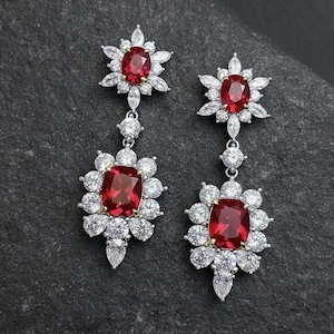 May include: A pair of elegant drop earrings. Each earring features a large, rectangular, deep red gemstone surrounded by a halo of smaller, clear gemstones. The earrings have a floral design with a smaller red gemstone at the top.