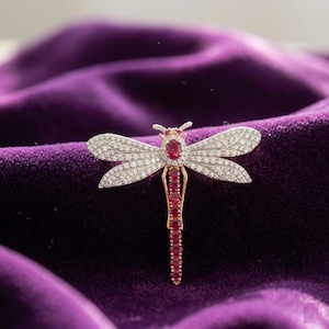 Puede incluir: Broche de libélula con cuerpo dorado, gemas de color rubí y alas adornadas con pequeños cristales transparentes. El broche se encuentra sobre un fondo de terciopelo morado oscuro, resaltando su diseño detallado y sus detalles brillantes.