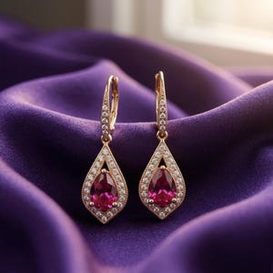 May include: A pair of gold-colored drop earrings with a teardrop-shaped, deep pink gemstone center. The gemstones are surrounded by small, clear crystals. The earrings are displayed on a purple satin fabric.