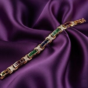 May include: A gold-toned bracelet with rectangular gemstones in green, blue, and burgundy, interspersed with diamond accents. The bracelet is displayed on a purple satin fabric, creating a luxurious aesthetic.