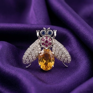 Puede incluir: Broche de abeja plateada con un cuerpo de gema amarilla ovalada, detalles de rubíes y alas con diamantes. El broche tiene ojos de gema azul y está sobre un fondo de satén púrpura.