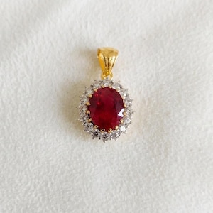 May include: A gold pendant with a large, oval, deep red gemstone, surrounded by a halo of smaller, clear stones. The pendant hangs from a gold bail. The jewelry piece is set against a white, textured background.