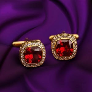 May include: A pair of gold cufflinks featuring large, square, red gemstones. Each cufflink is framed by a border of small, clear crystals. The cufflinks are presented on a deep purple, textured fabric.