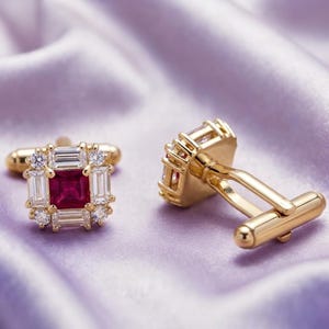 May include: A pair of gold cufflinks featuring a square-cut deep red center stone surrounded by a border of rectangular baguette-cut and round brilliant-cut clear stones. The cufflinks have a polished gold metal finish and a hinged toggle backing.